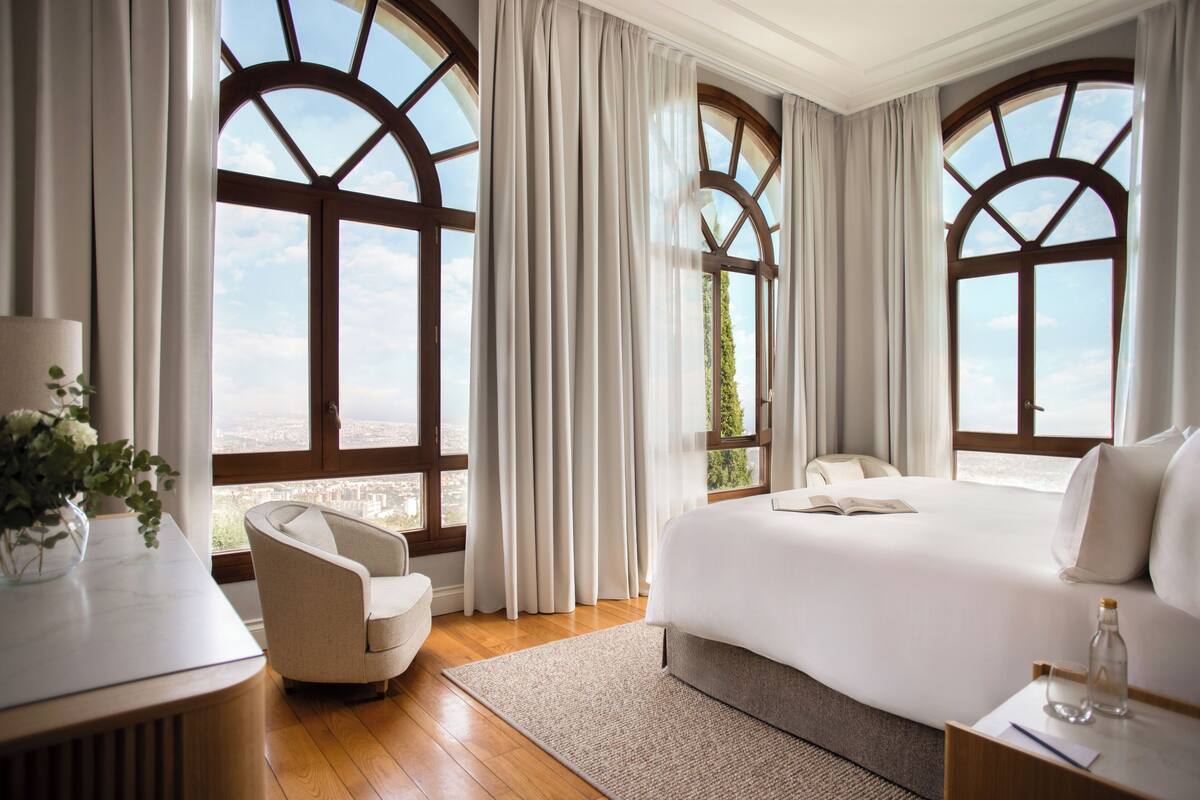 Quarto de hotel com uma cama grande, uma cadeira, uma escrivaninha e vista através de janelas em arco.