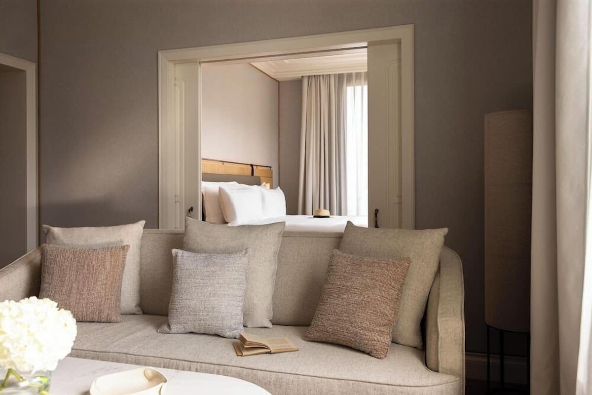 Quarto em tons neutros com sofá, um espelho refletindo uma cama e uma janela com cortinas.