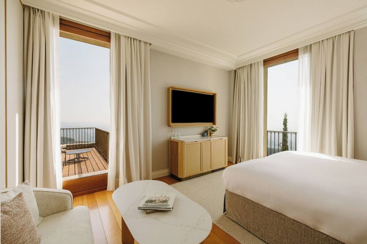 Quarto de hotel com uma cama grande, TV na parede, uma mesinha com uma revista e varanda com vista.