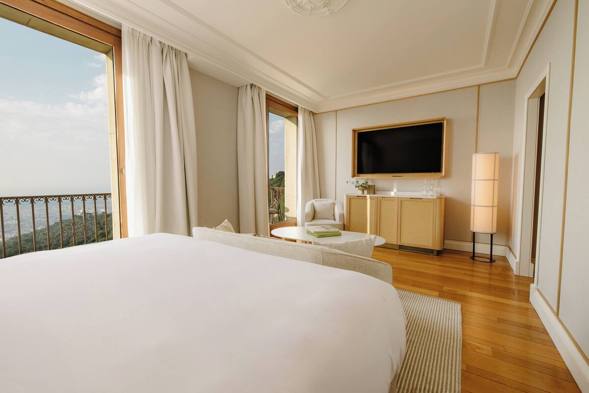 Quarto de hotel com uma cama grande, televisão na parede, mesinha de cabeceira com abajur e vista para o exterior.