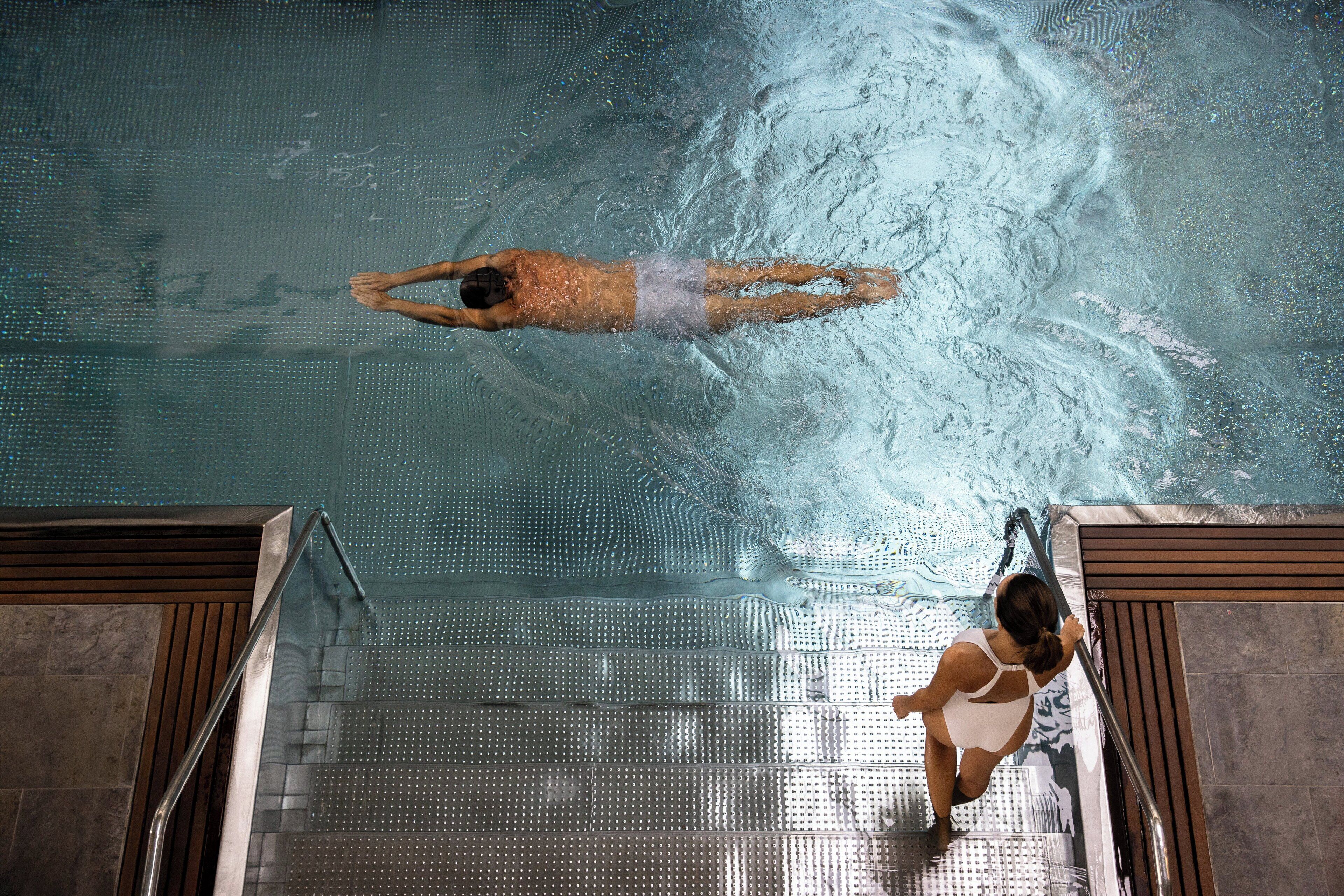A swimming pool with a person swimming and another person standing at the pool's edge.
