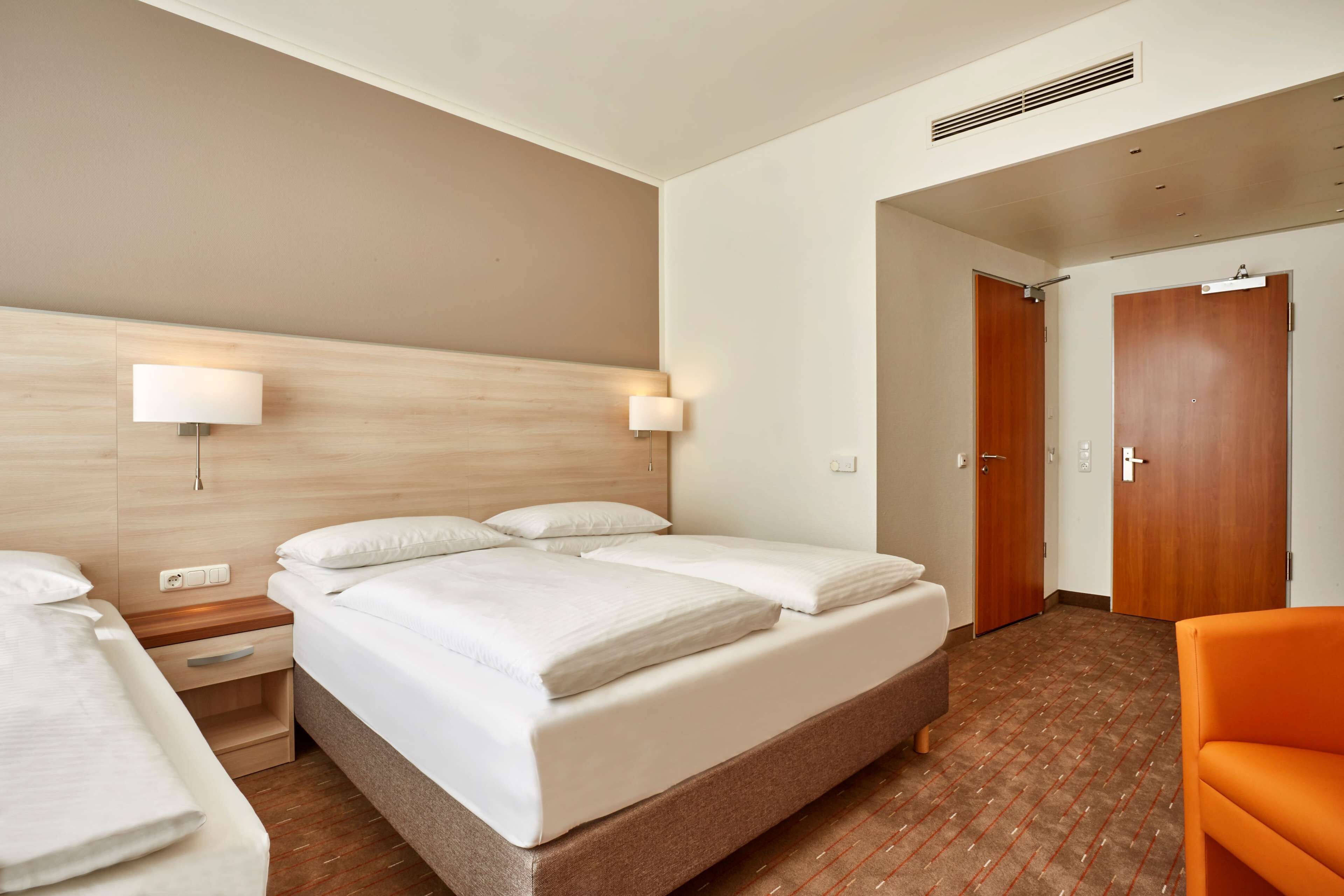 Ein Hotelzimmer mit einem großen Bett, Nachttischen, einem Beistelltisch, einem orangefarbenen Sessel und einem Holz-Kopfteil.