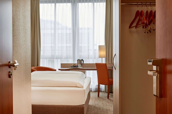A hotel room with a bed, a desk, an orange chair, a lamp, and a coat rack with hangers. - H+ Hotel Berlin Mitte (Berlin)