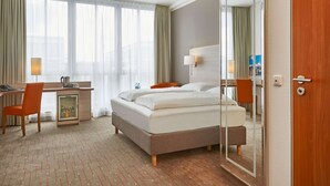 A hotel room with a bed, a desk with a chair, a small refrigerator, and a large mirror. - H+ Hotel Berlin Mitte (Berlin)