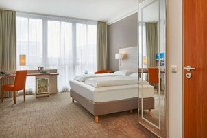 A hotel room with a bed, a desk with a chair, a small refrigerator, and a large mirror.