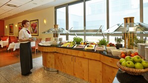 A buffet area with a variety of food items, a waiter, and a view of the city through large windows. - H+ Hotel Berlin Mitte (Berlin)