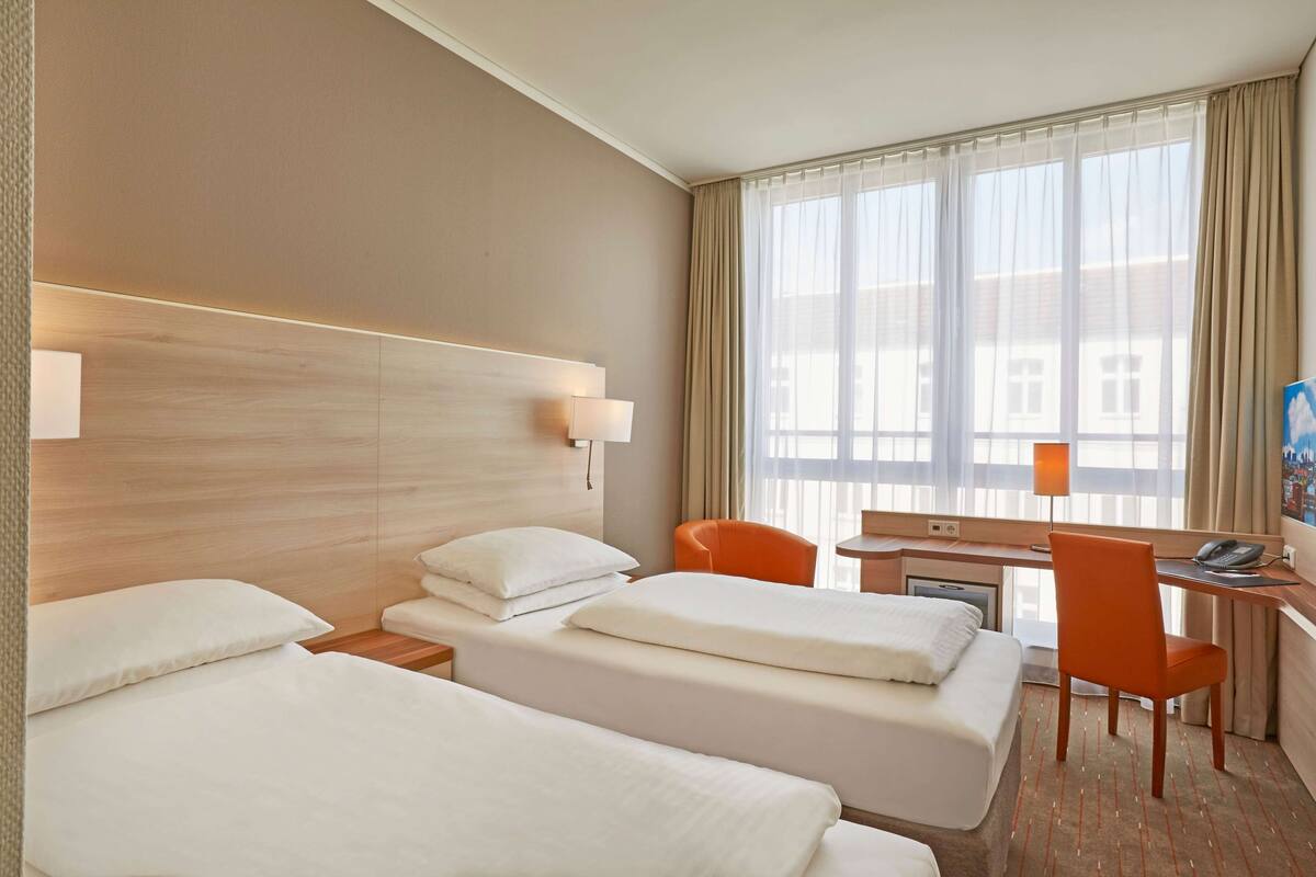 A hotel room with two beds, a desk, an orange chair, and a large window with curtains.