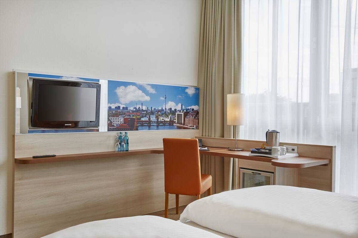 A hotel room with a bed, a desk with a computer, a chair, a TV, and a cityscape painting on the wall.