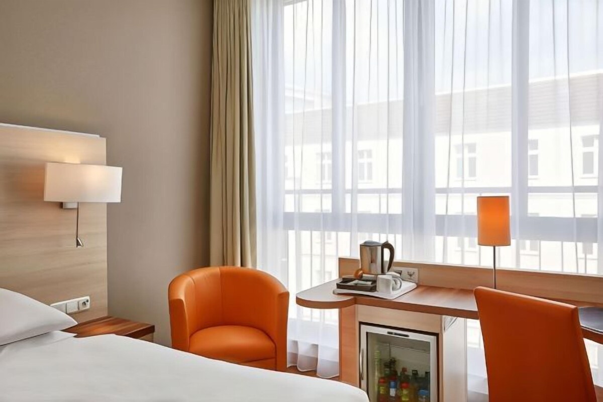 Une chambre d’hôtel avec un lit, deux fauteuils orange, un bureau avec une machine à café, un mini-réfrigérateur et une grande fenêtre avec des rideaux.