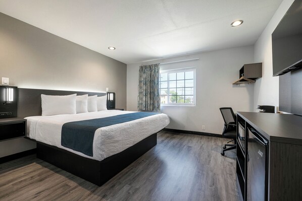A modern hotel room with a large bed, a desk, a chair, a window with curtains, and a flat-screen TV.
