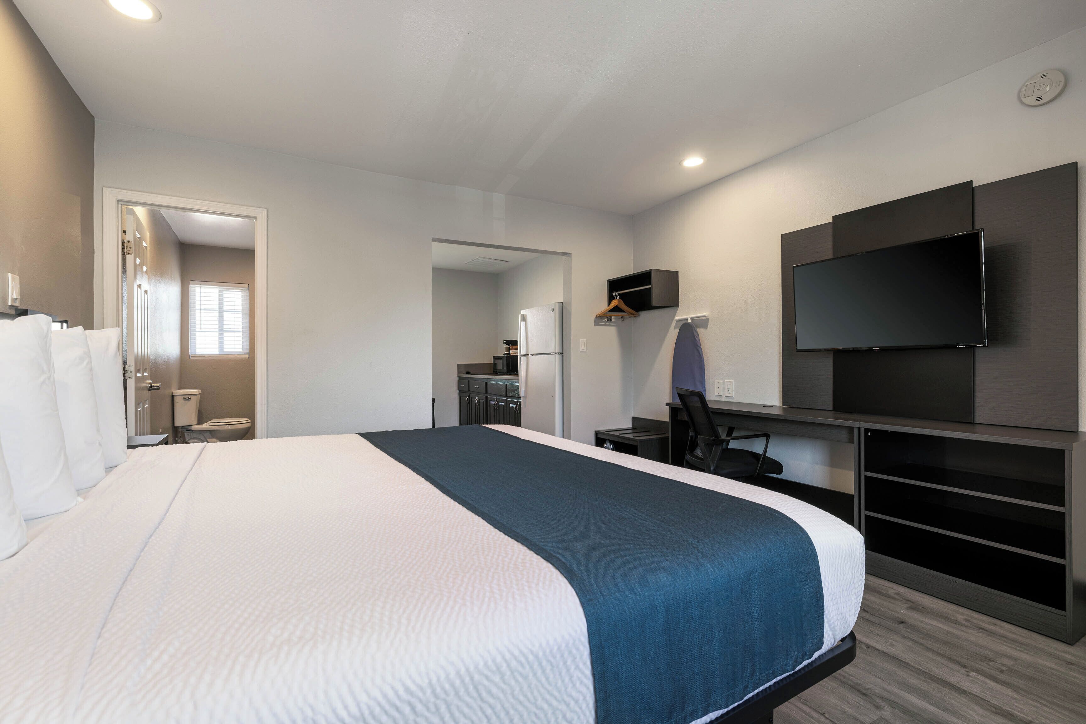 A hotel room with a large bed, a desk with a chair, a television, and a kitchenette in the background.
