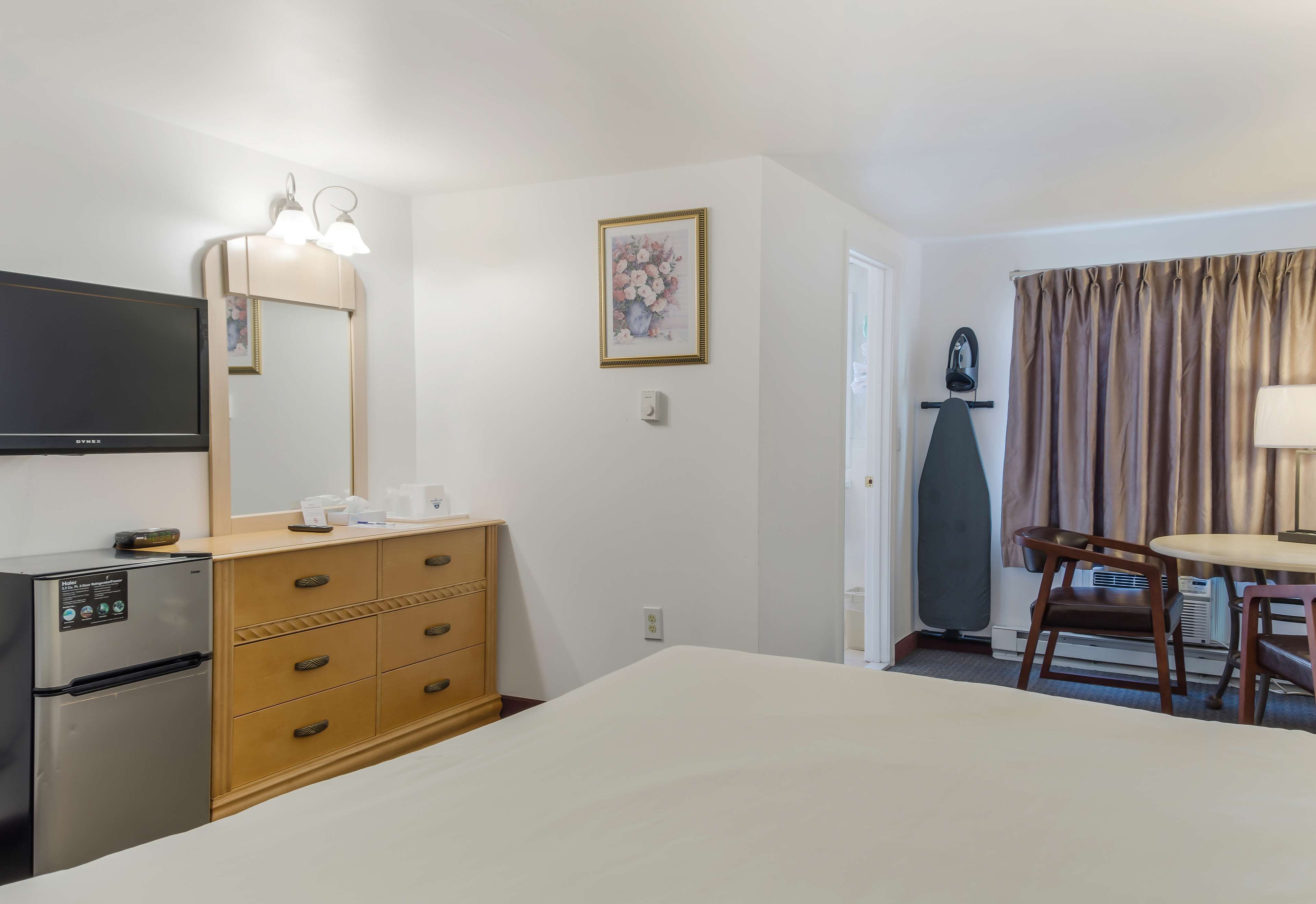 A hotel room with a bed, a TV, a minibar, a wooden dresser, a desk with a chair, and a door leading to the bathroom.