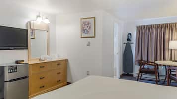 A hotel room with a bed, a TV, a minibar, a wooden dresser, a desk with a chair, and a door leading to the bathroom.