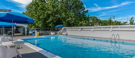 A rectangular swimming pool with clear blue water, surrounded by white lounge chairs and a blue umbrella.