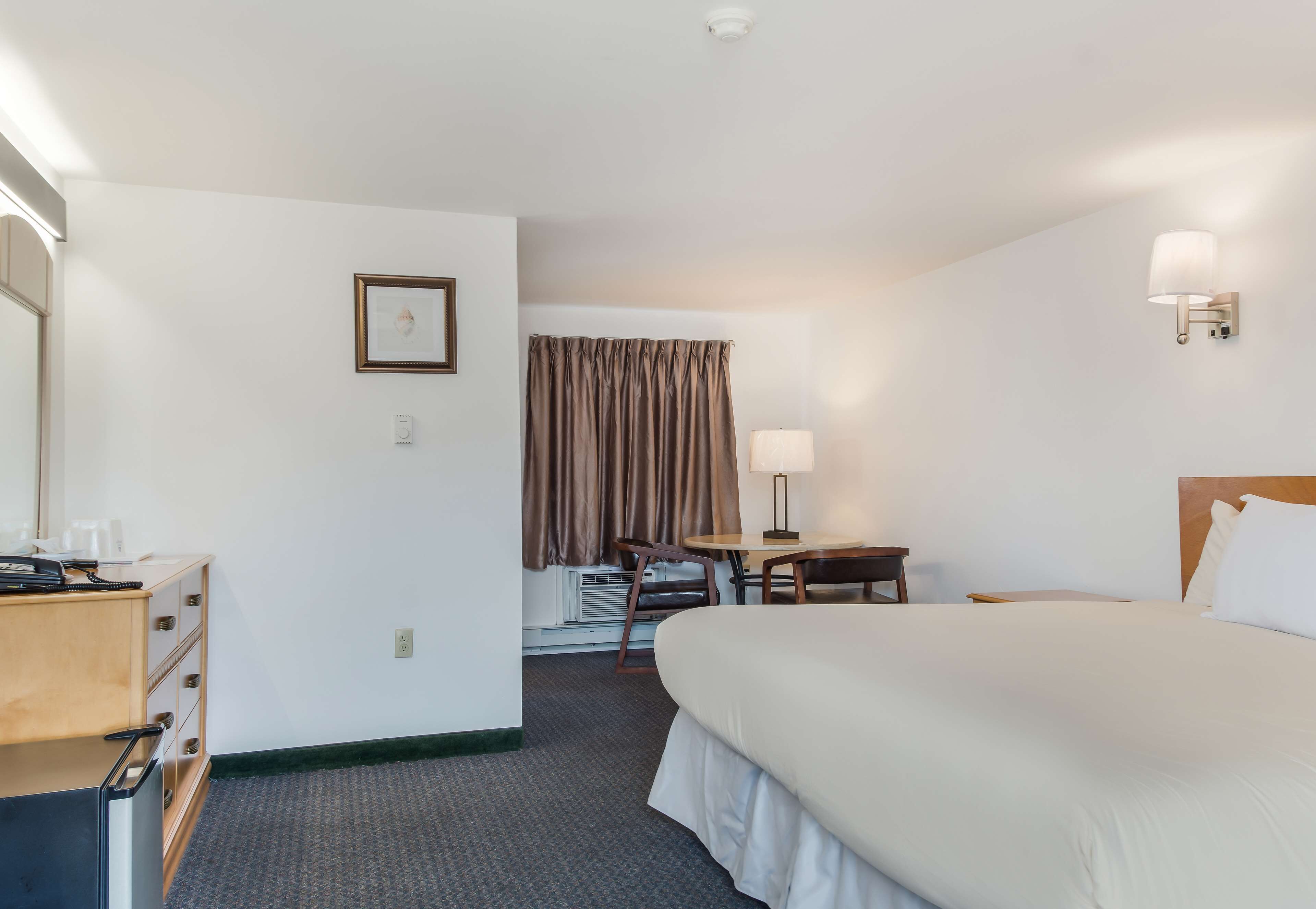 A hotel room with a large bed, a desk with a chair, a small table, a lamp, a picture on the wall, and a window with curtains.
