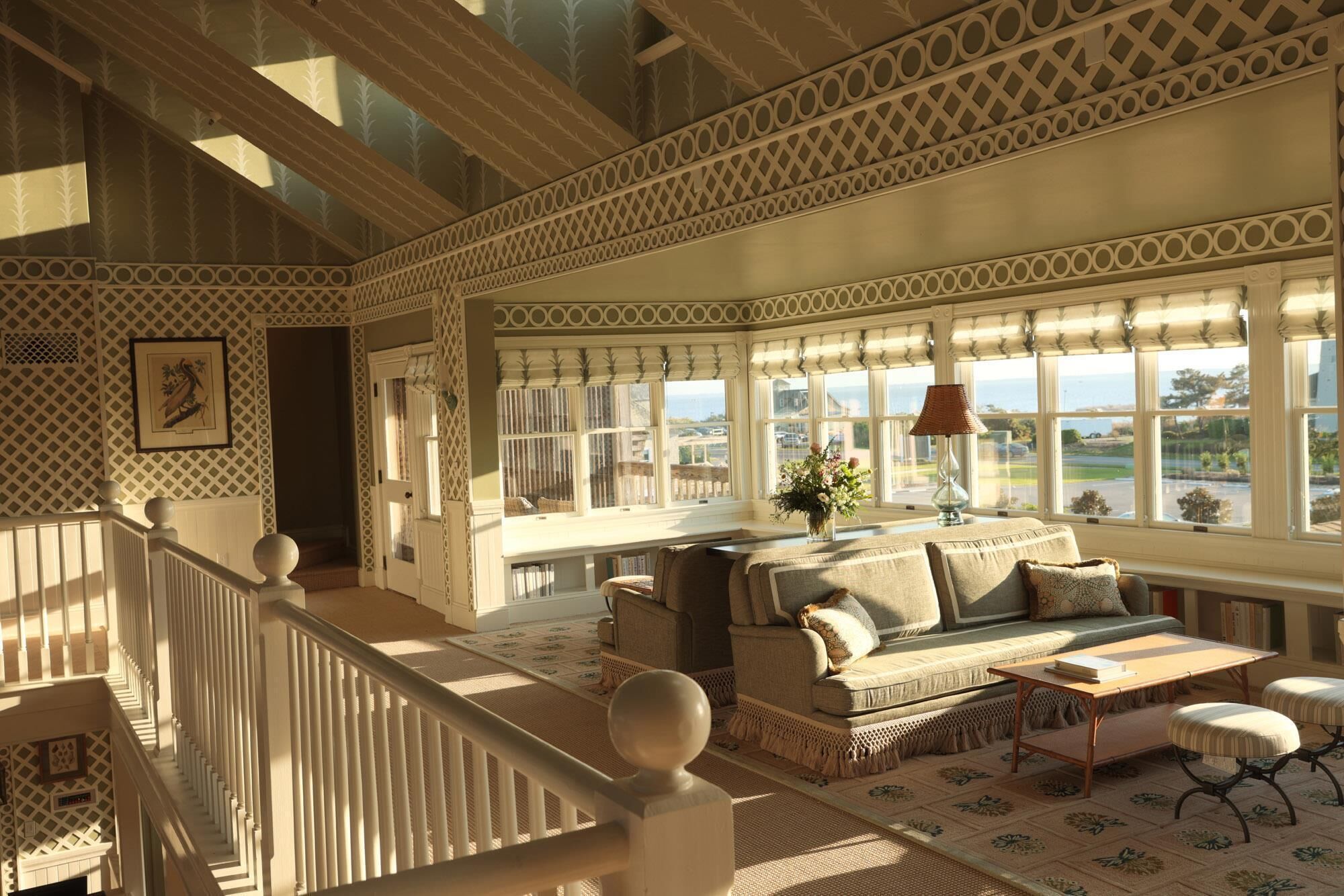 A spacious room with a high ceiling, patterned walls, a large window with a view, and a cozy seating area with a sofa and armchairs.