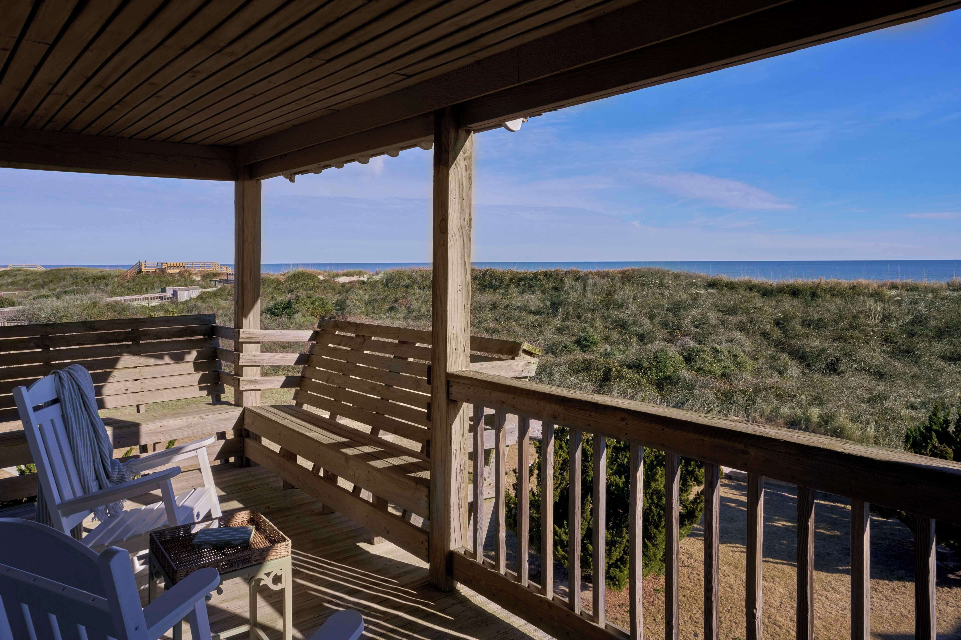 A wooden deck with two white chairs and a basket, overlooking a beach and dunes.