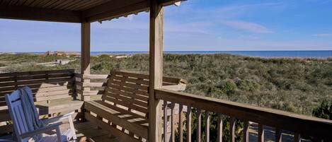 A wooden deck with two white chairs and a basket, overlooking a beach and dunes.