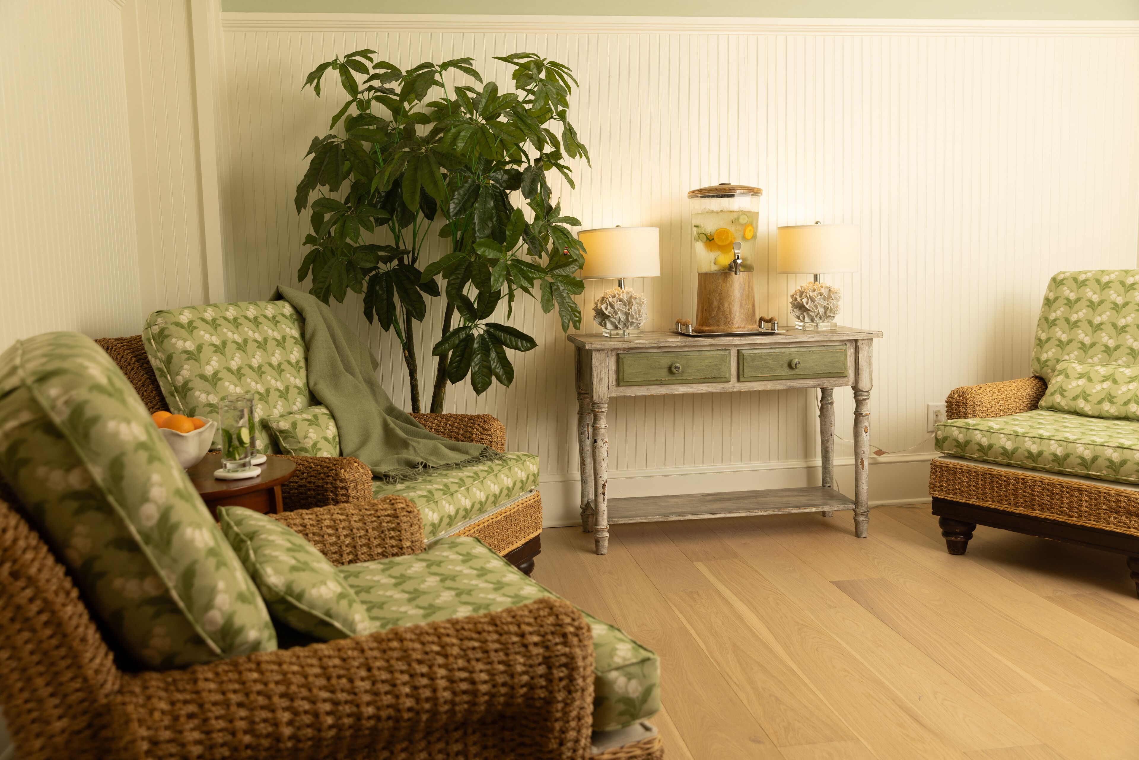 A cozy living room with wicker furniture, a large plant, and a side table with lamps and a decorative vase.