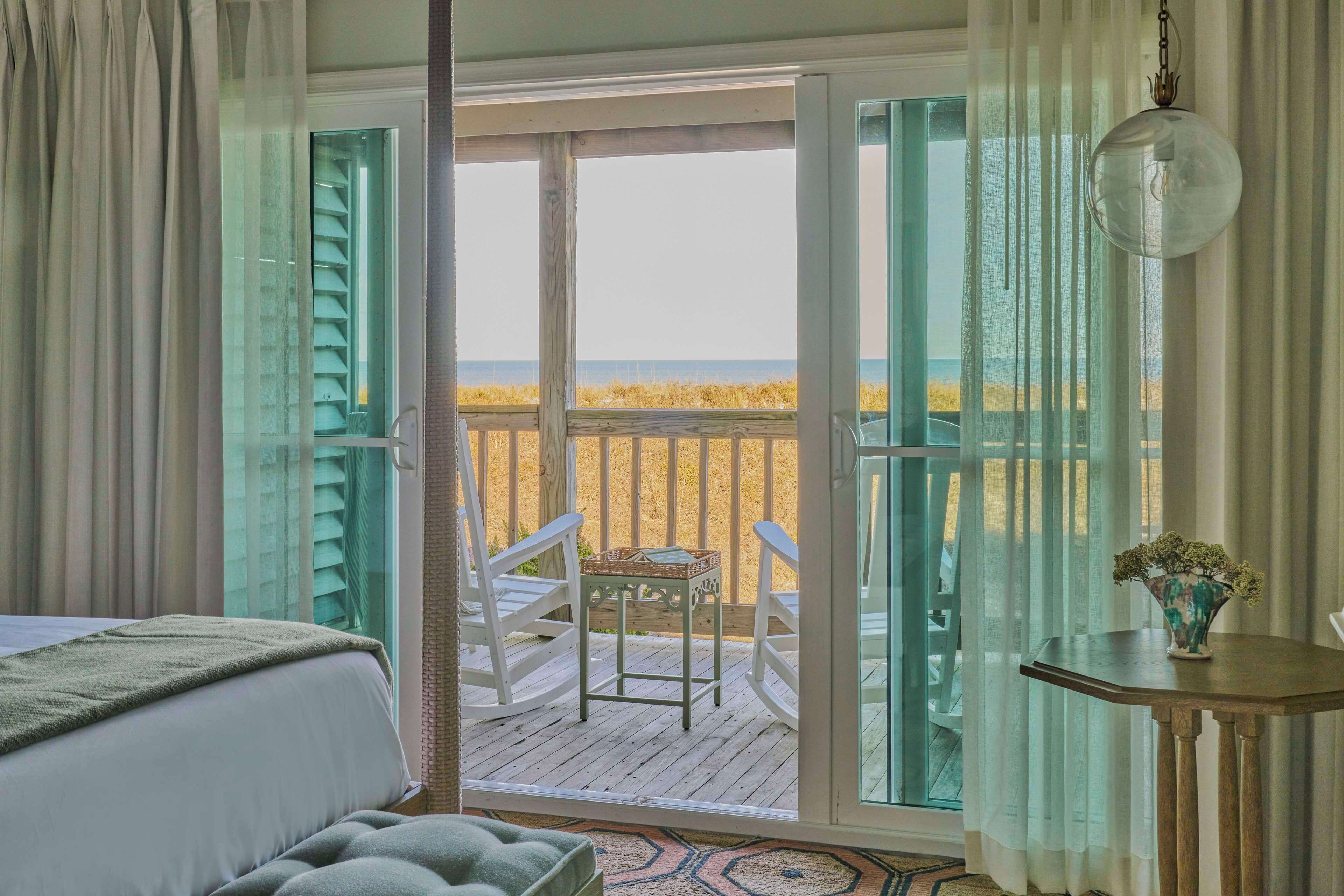 A bedroom with a view of the beach through open French doors, a bed, a small table with a vase, and a chair on a wooden deck.