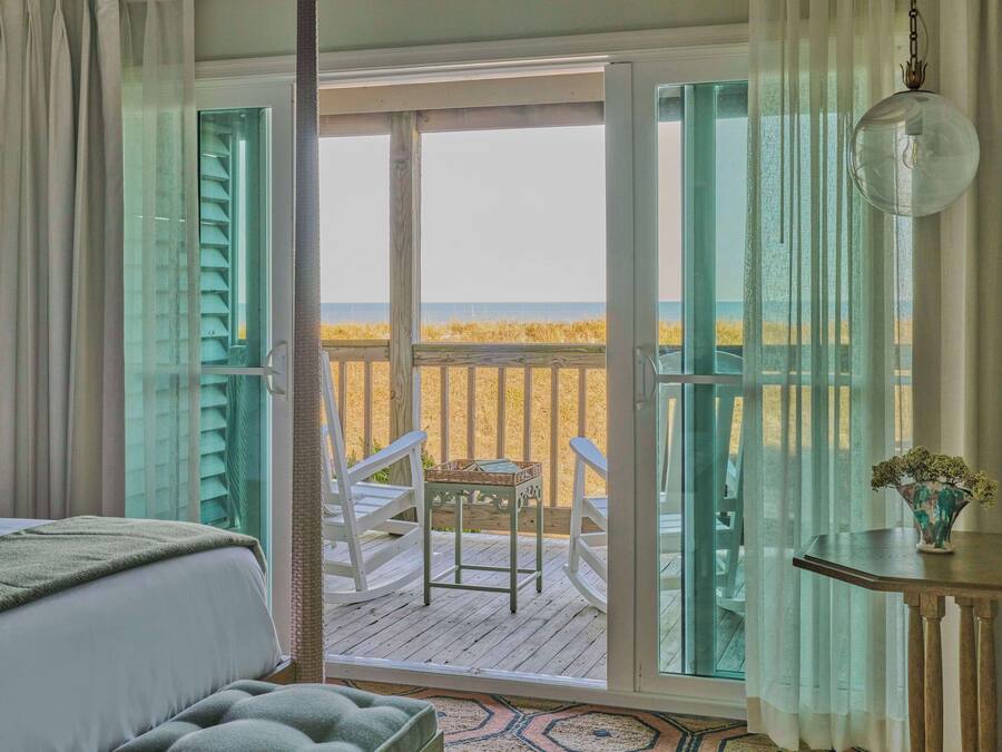 A bedroom with a view of the beach through open French doors, a bed, a small table with a vase, and a chair on a wooden deck.