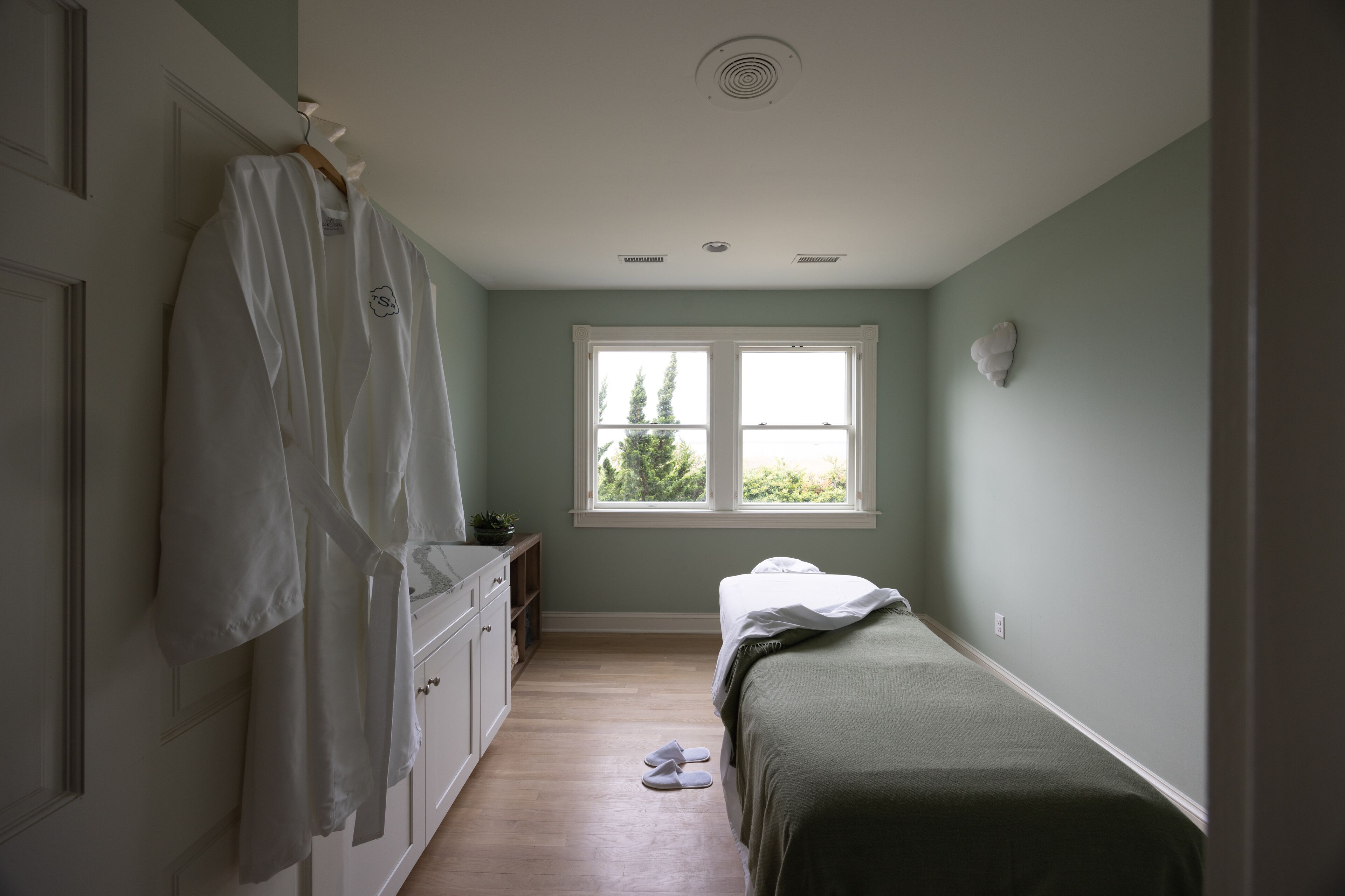 A bedroom with a single bed, a window, a wardrobe, and a pair of slippers on the floor.