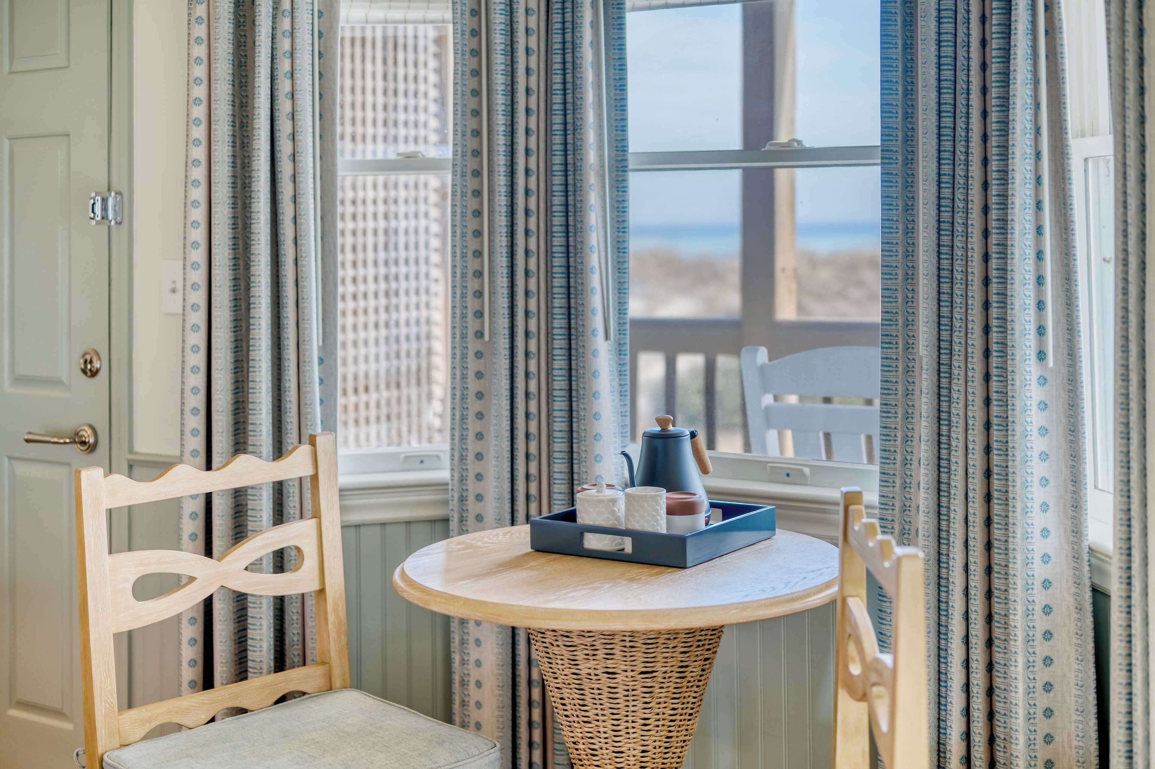 A room with a round table, wicker chair, and a view of the beach through a window with patterned curtains.
