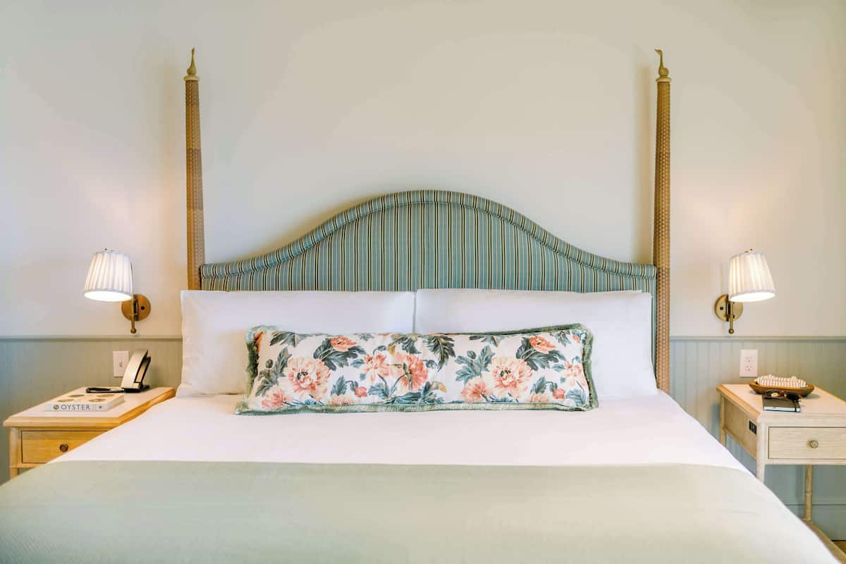 Un lit bien fait, agrémenté d’un coussin à motifs floraux, de deux lampes de chevet et d’une table de nuit sur laquelle se trouvent un livre et un téléphone.