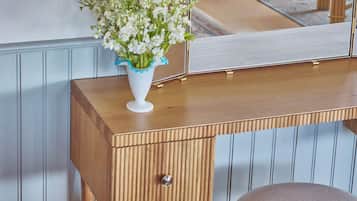 A wooden vanity with a mirror, a white vase with flowers, and a wooden stool.