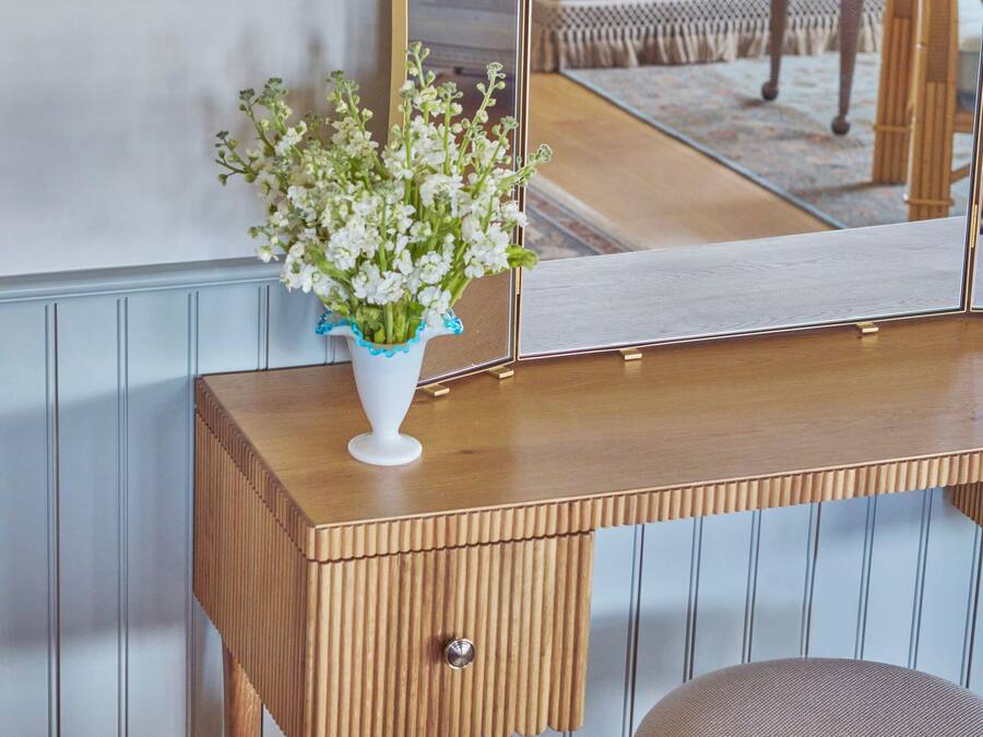 A wooden vanity with a mirror, a white vase with flowers, and a wooden stool.