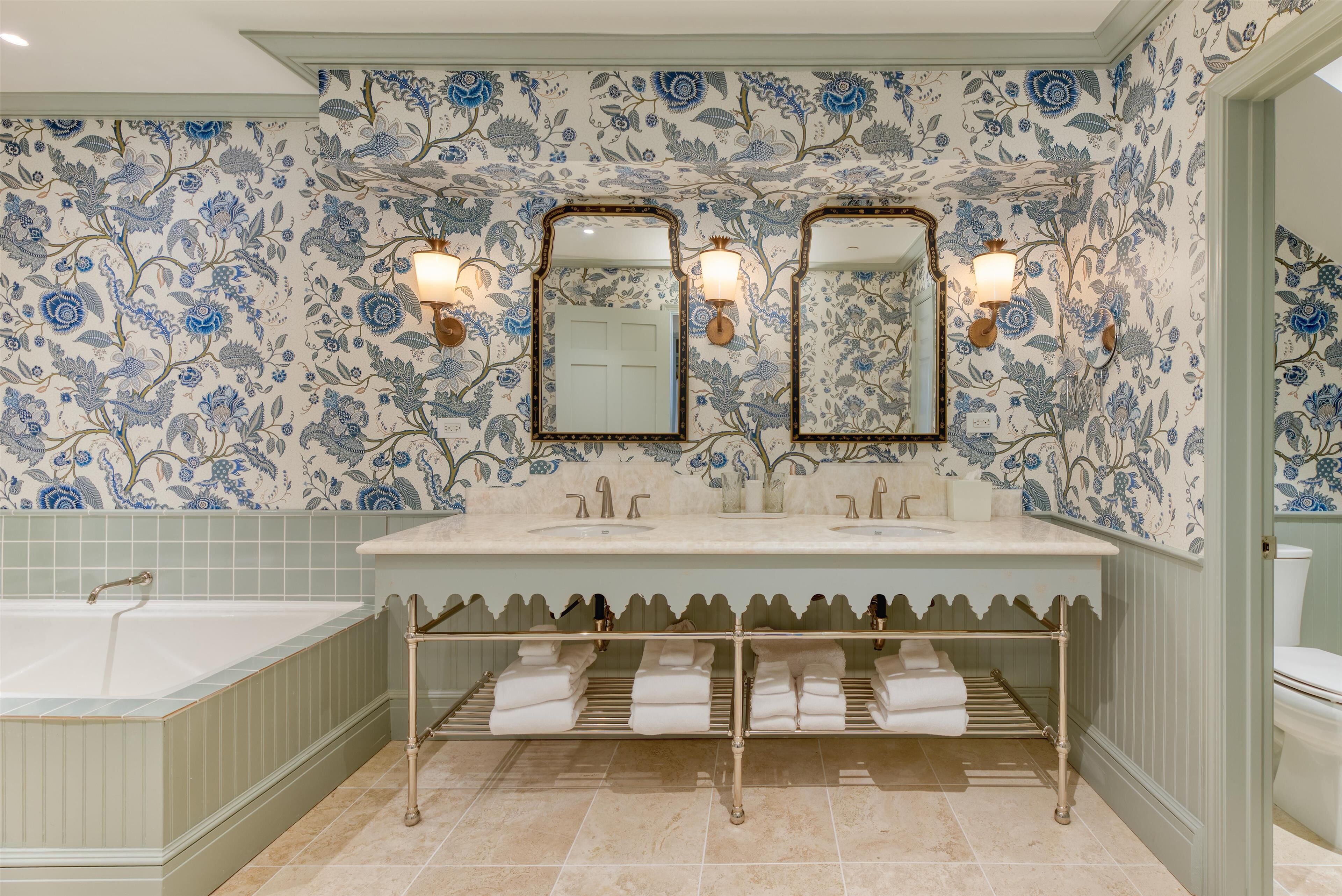 Une salle de bain avec un papier peint à motifs floraux, un meuble-lavabo à deux vasques, une baignoire et un porte-serviettes.