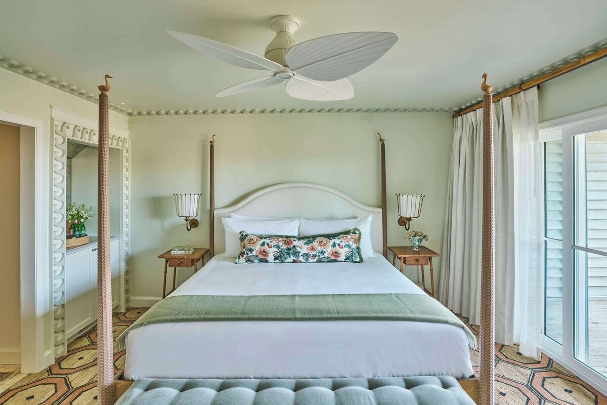 Un lit à baldaquin avec une tête de lit blanche, deux tables de chevet avec des lampes, une grande fenêtre avec des rideaux et un tapis à motifs.
