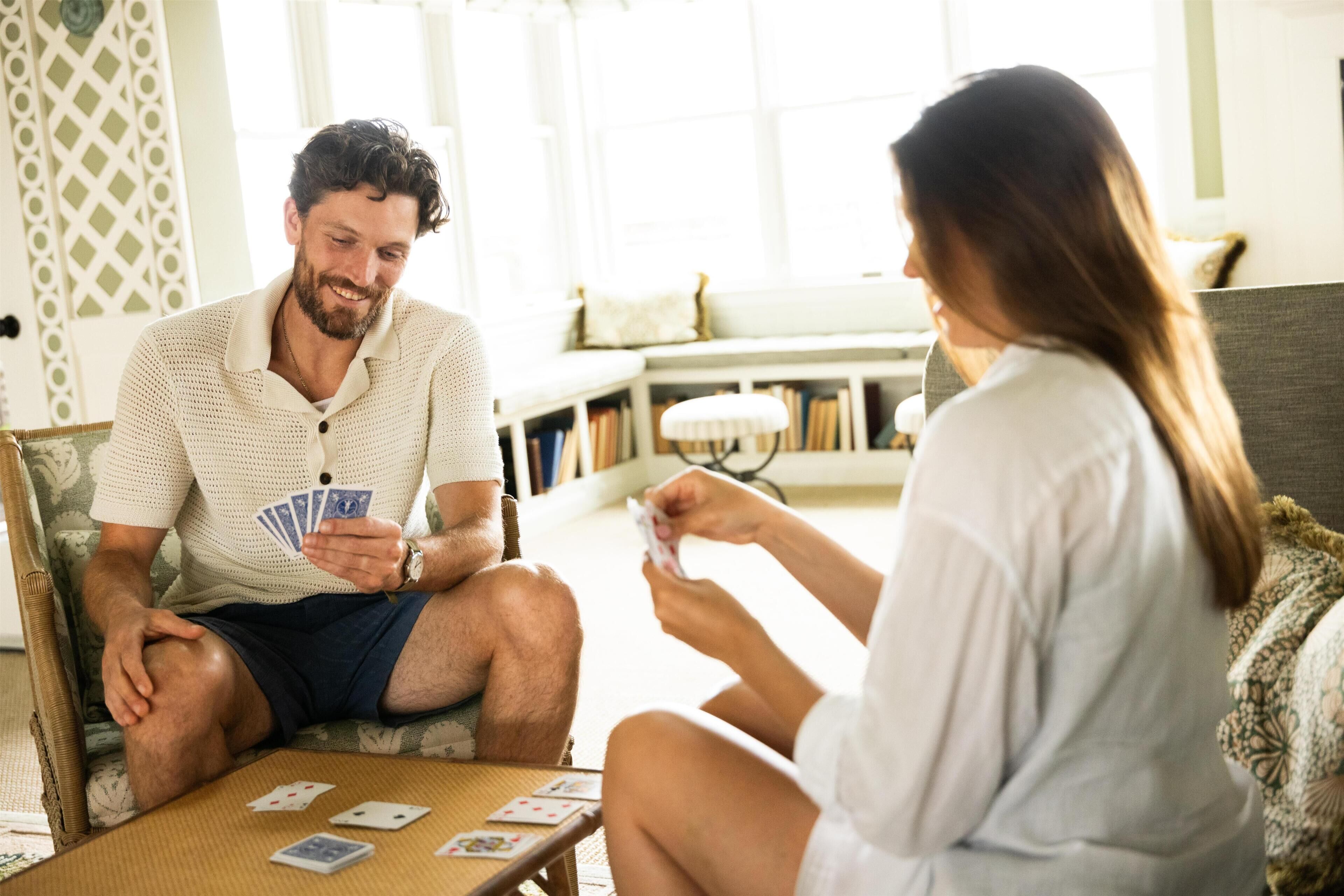 Two people playing cards on a couch in a well-lit room with a bookshelf and a patterned wall.