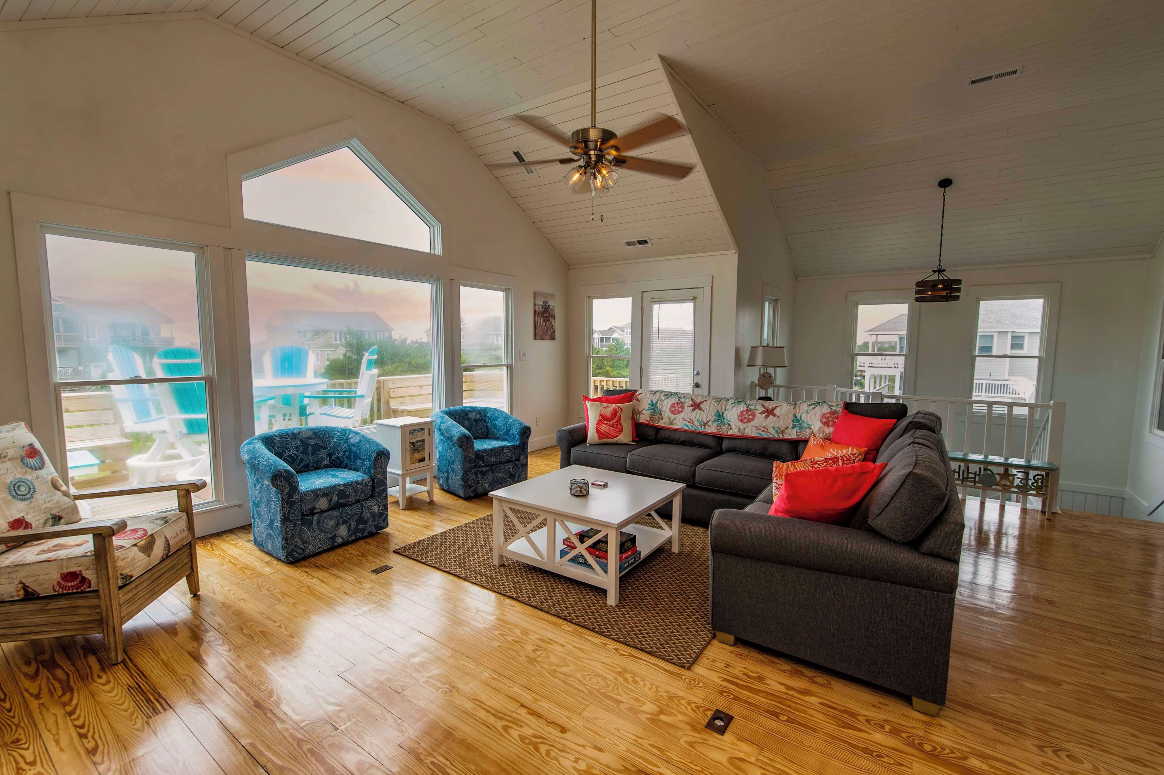 A spacious living room with a high ceiling, large windows, a ceiling fan, and a view of a residential area.
