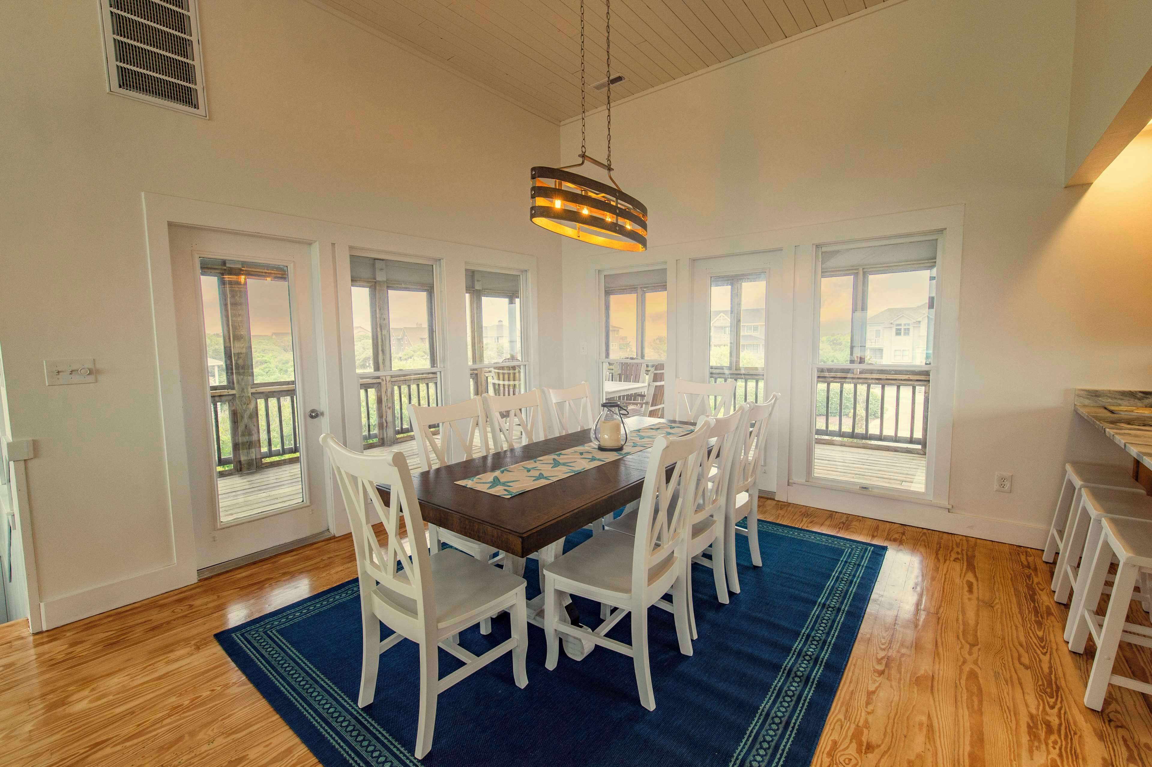 Un coin repas avec une table en bois, des chaises blanches, un tapis bleu et de grandes fenêtres.