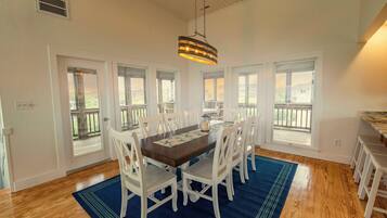Un coin repas avec une table en bois, des chaises blanches, un tapis bleu et de grandes fenêtres.