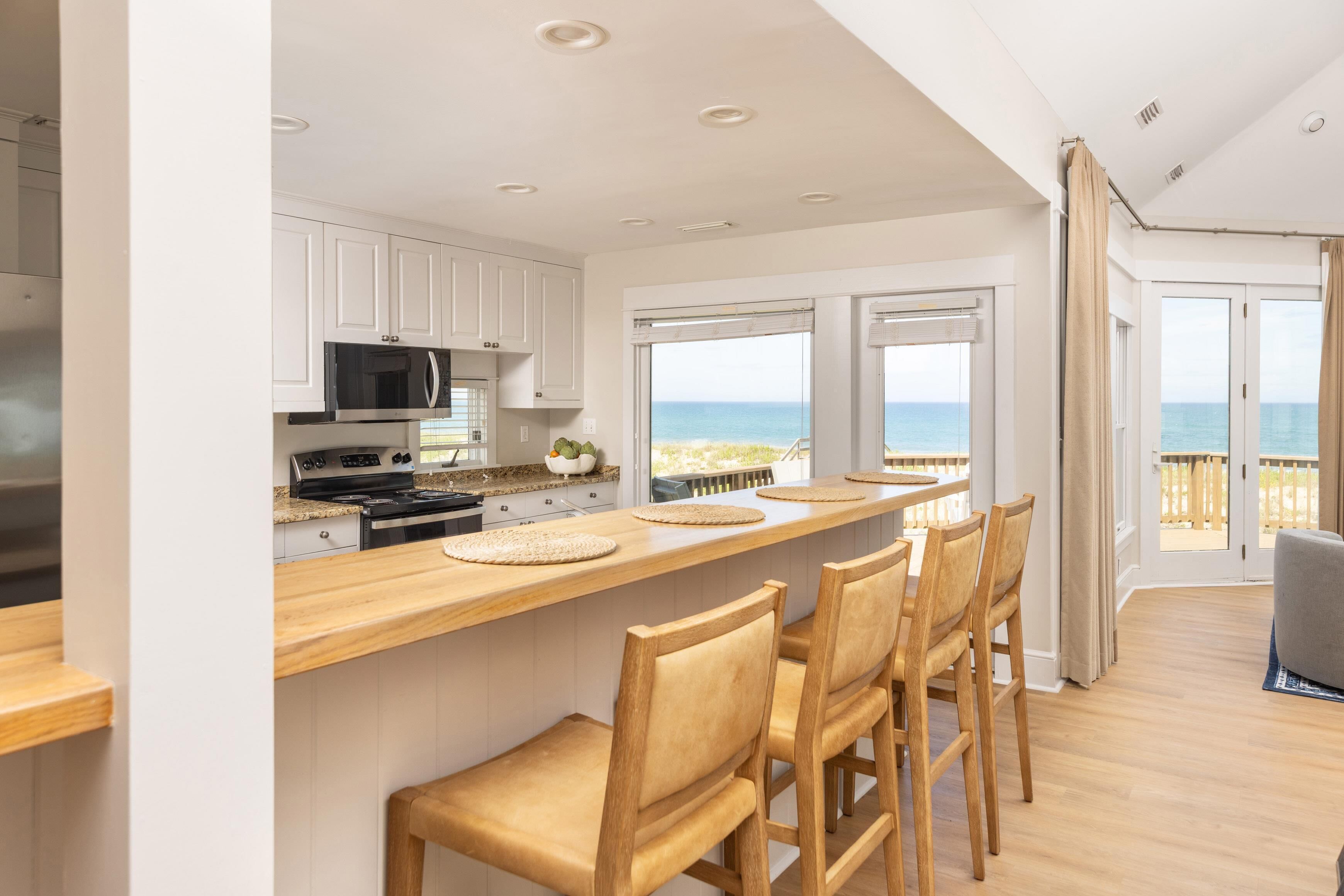 A modern kitchen with white cabinets, a wooden countertop, and a view of the ocean through large windows.