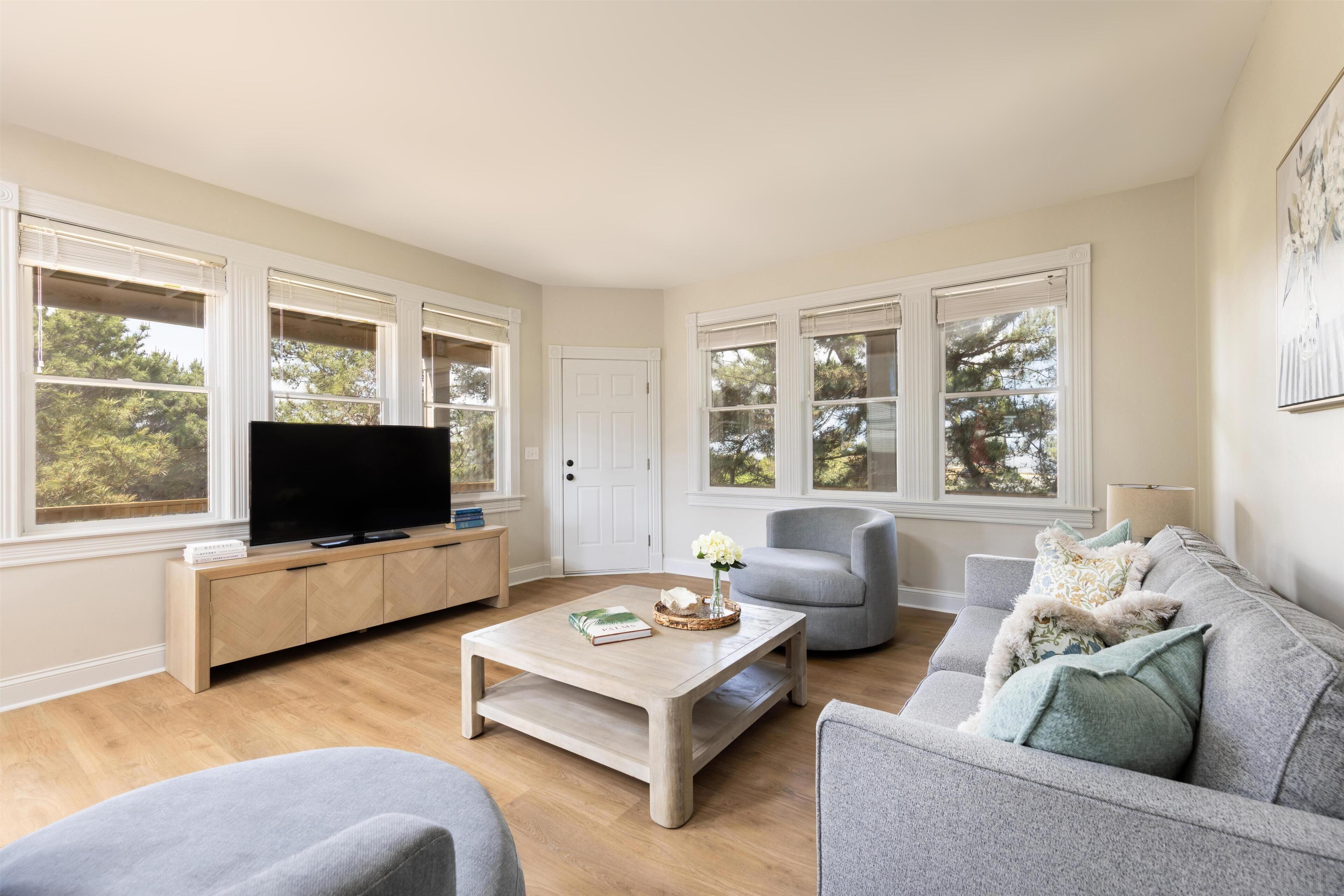 A living room with a television on a wooden cabinet, a grey sofa, and a coffee table with a book and vase.
