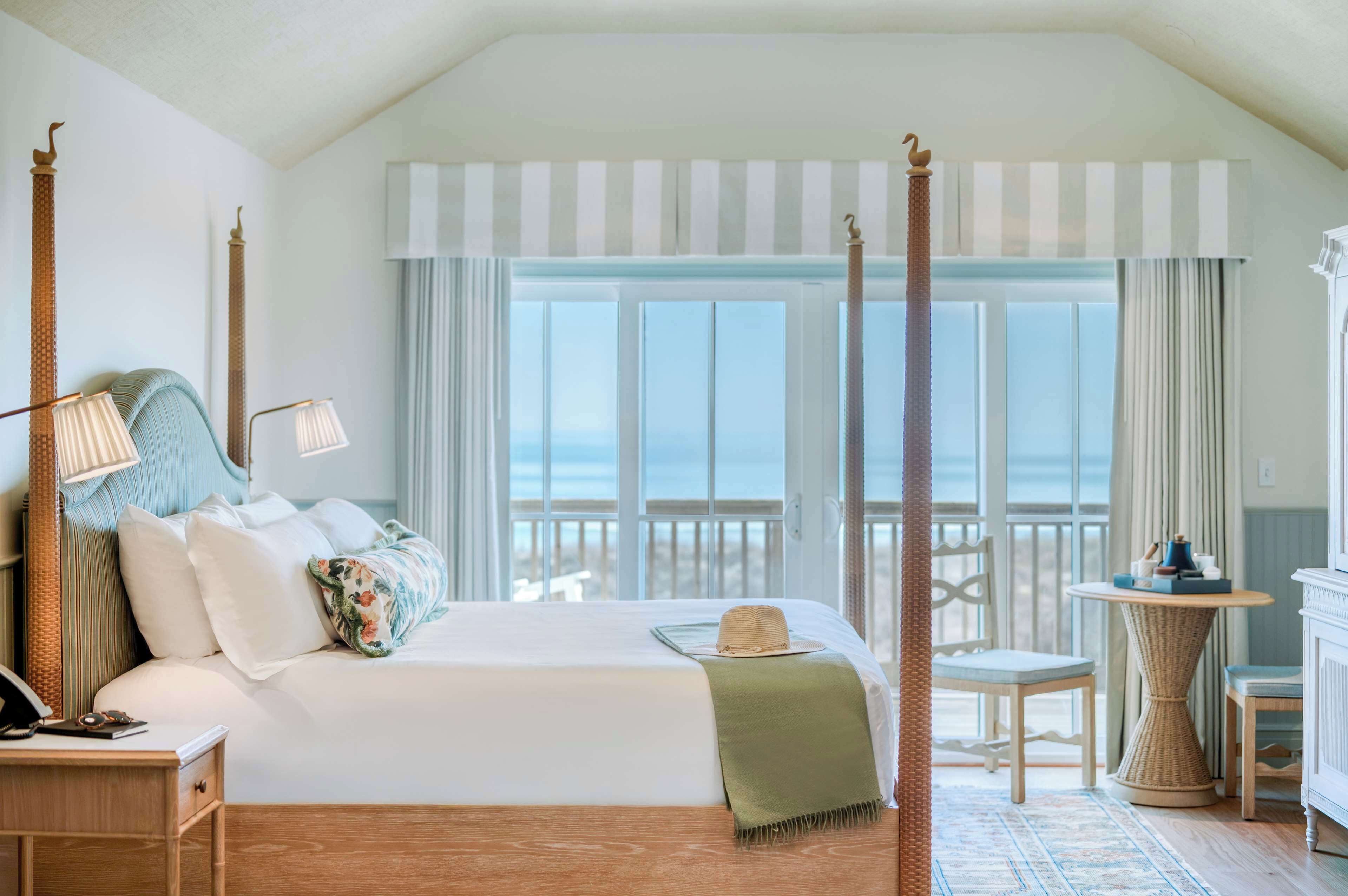 Una cama con dosel y vistas al océano, una mesita con una lámpara, y una silla junto a la ventana.