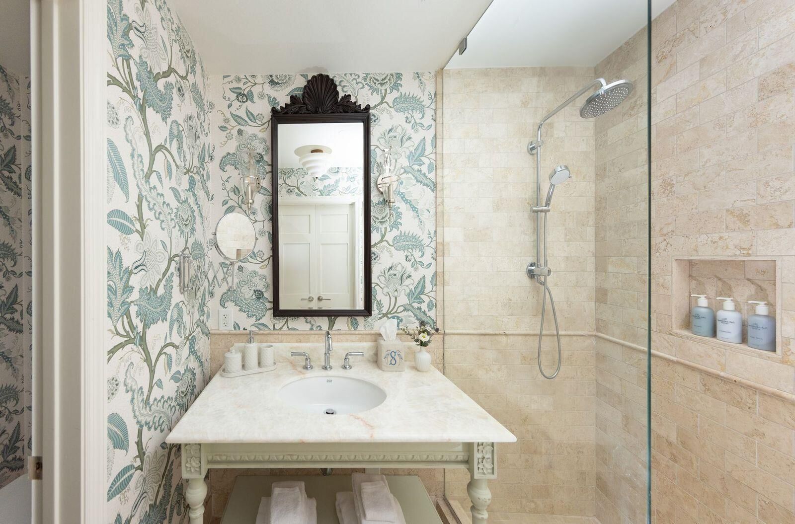 A bathroom with a marble sink, a large mirror, a shower, and a shelf with toiletries.