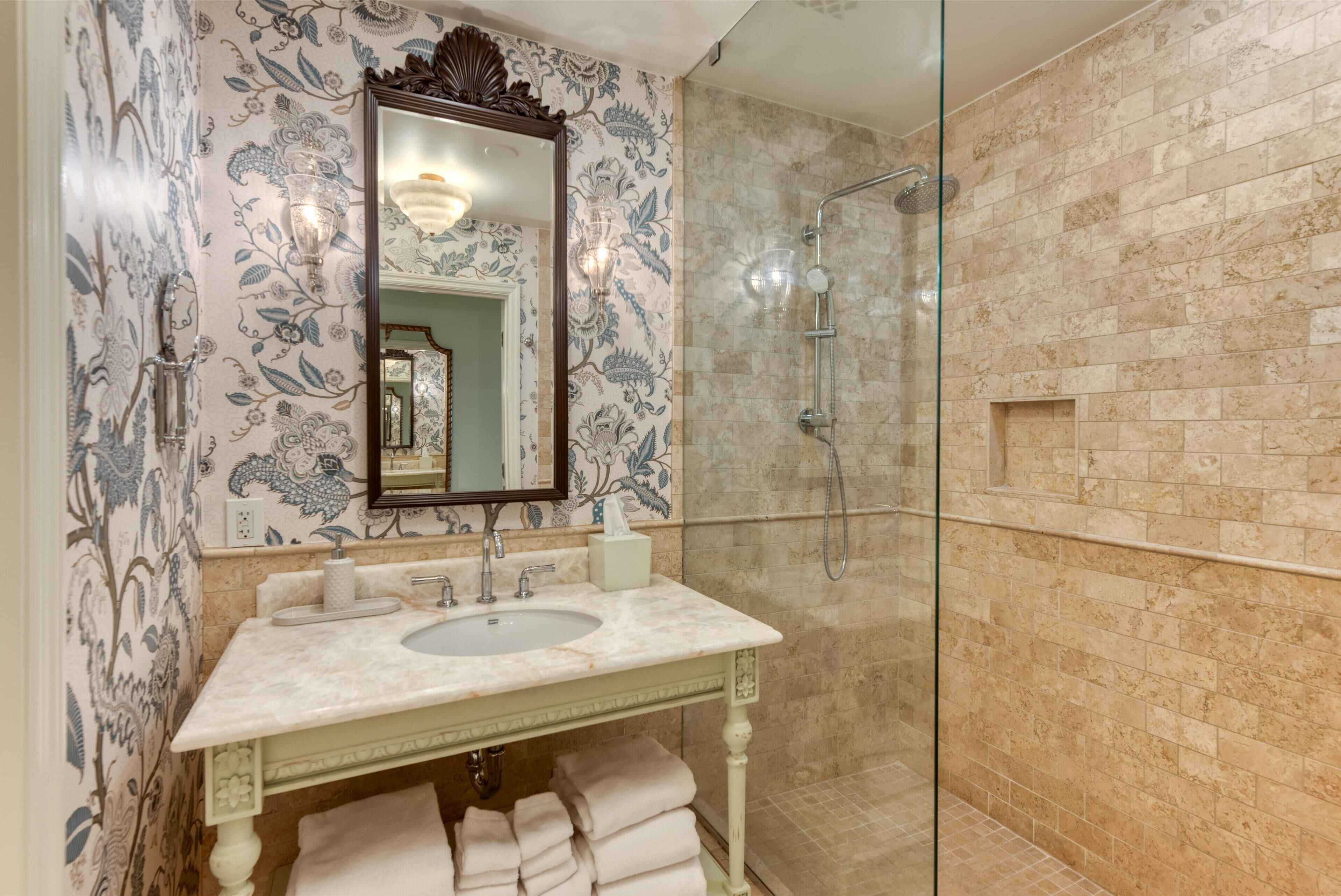 Un baño con lavamanos de mármol, cabina de ducha de vidrio y papel tapiz estampado.