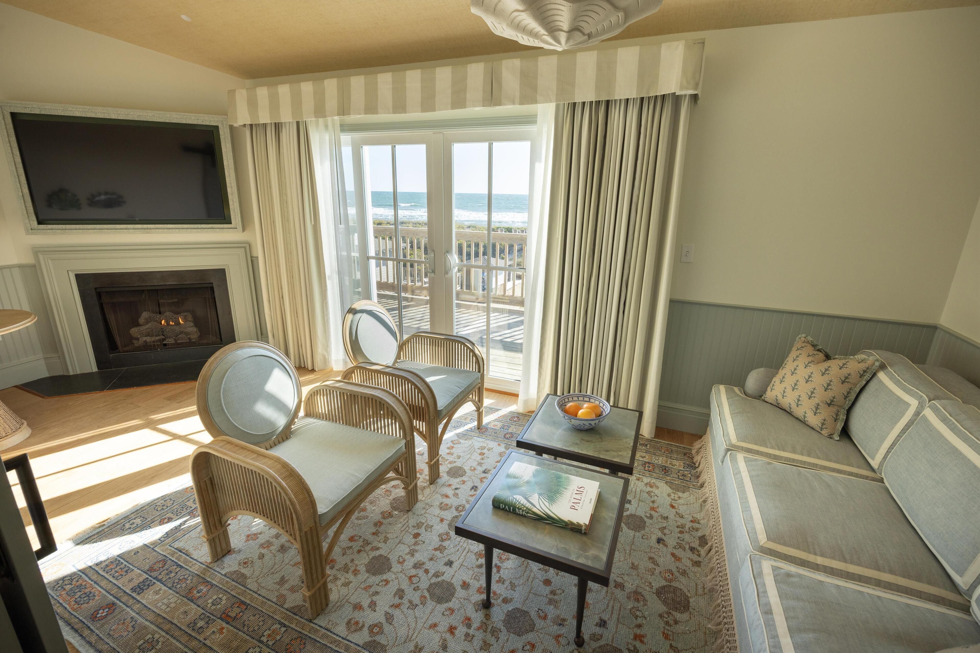 A living room with a fireplace, wicker chairs, a sofa, a coffee table, and a view of the ocean through large windows.