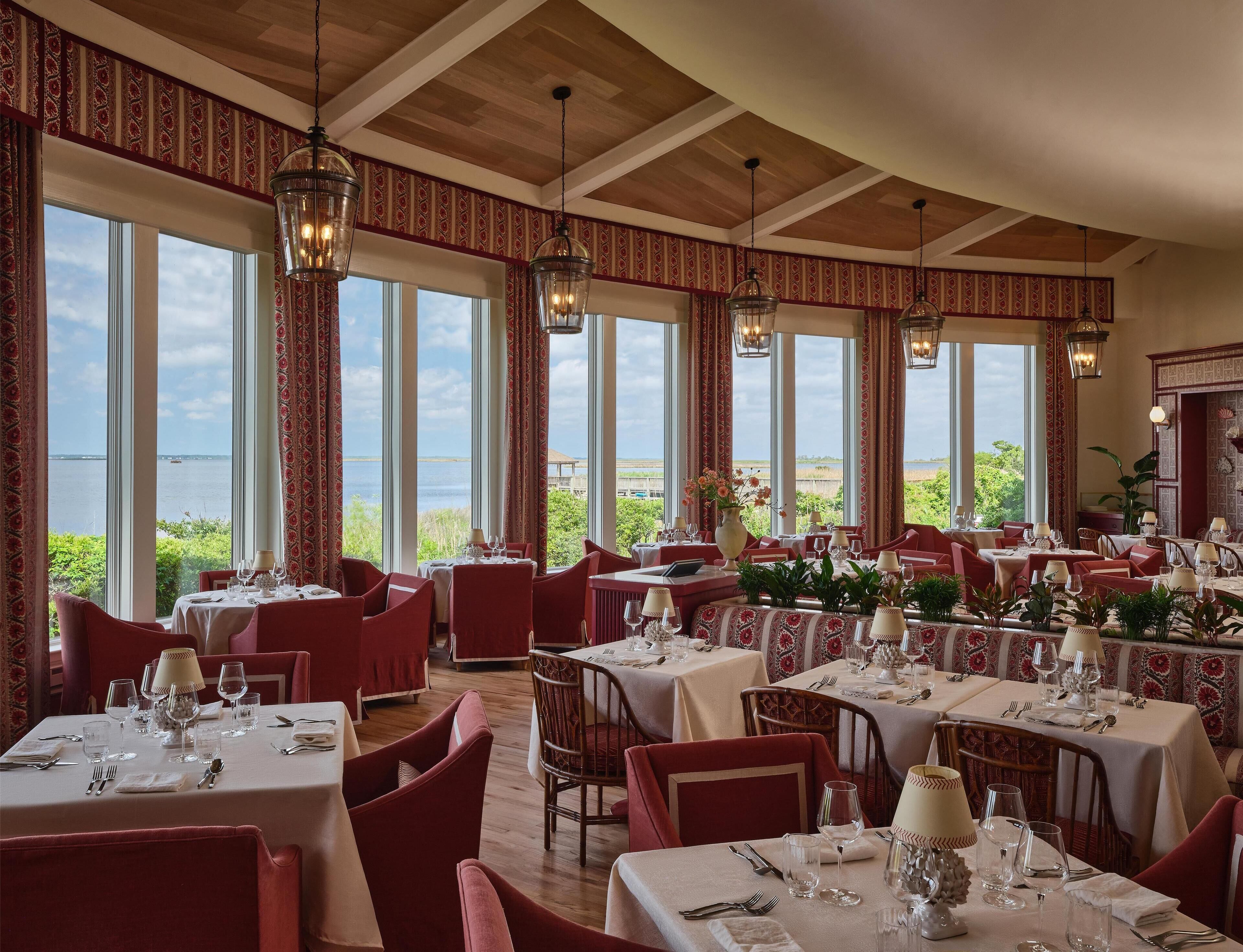 Een restaurant met meerdere tafels en stoelen, grote ramen met uitzicht op zee en hangende lantaarns.