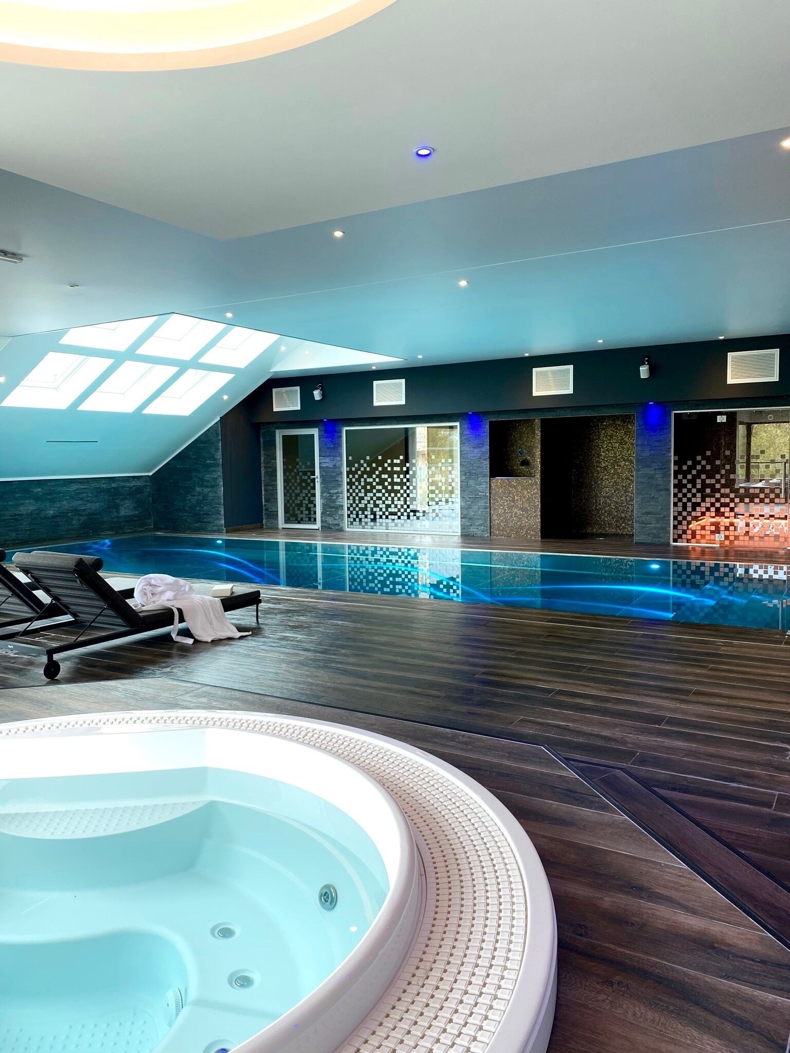 A modern indoor swimming pool with a hot tub, lounge chairs, and a skylight.
