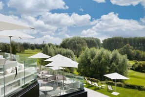 A modern outdoor seating area with glass railings, white umbrellas, and a view of a well-manicured lawn and trees. - Best Western Plus Le Fairway Hotel & Spa Golf d'Arras (Anzin-Saint-Aubin)