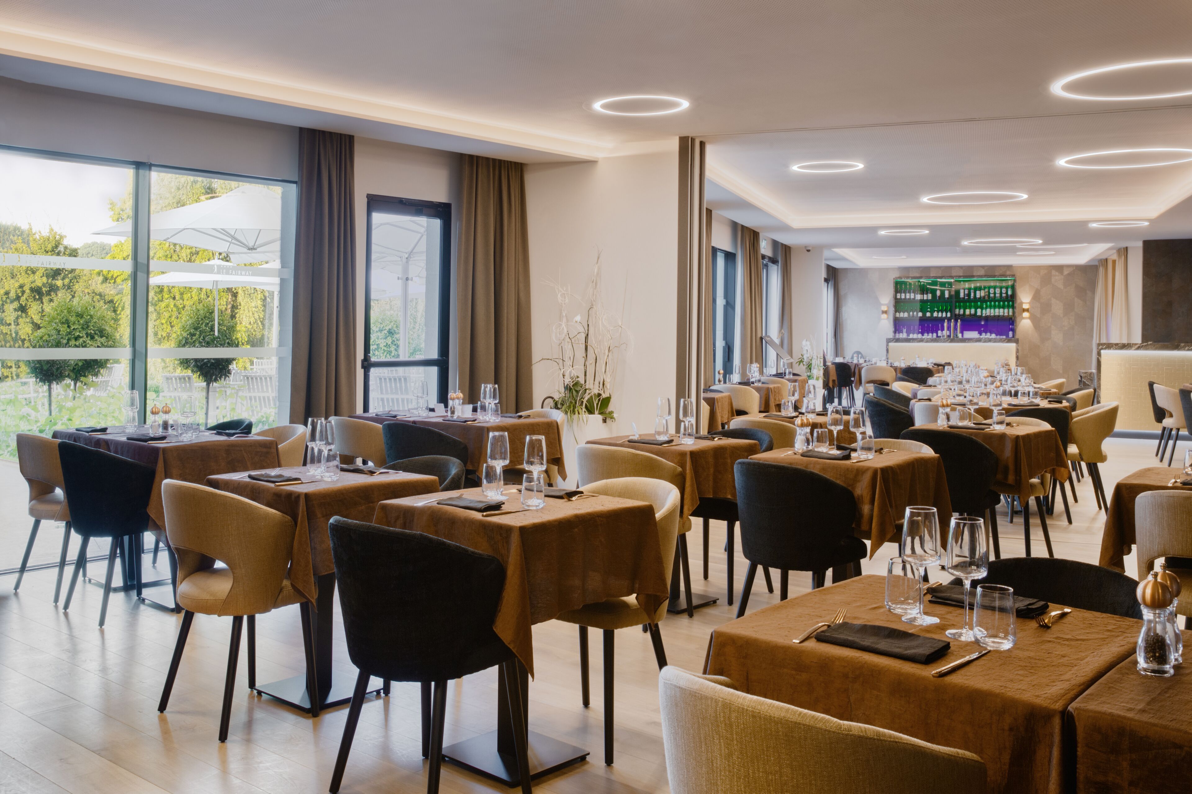 Een modern restaurant met meerdere gedekte tafels, grote ramen met uitzicht op groen en een bar op de achtergrond.