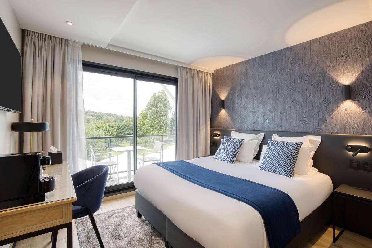 A modern hotel room with a large bed, a desk with a computer, and a view of the outdoors.