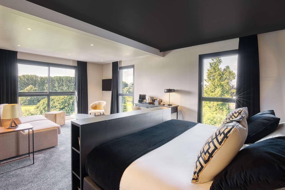 A modern hotel room with a large bed, a desk, a chair, and a sofa. There are large windows with a view of trees.