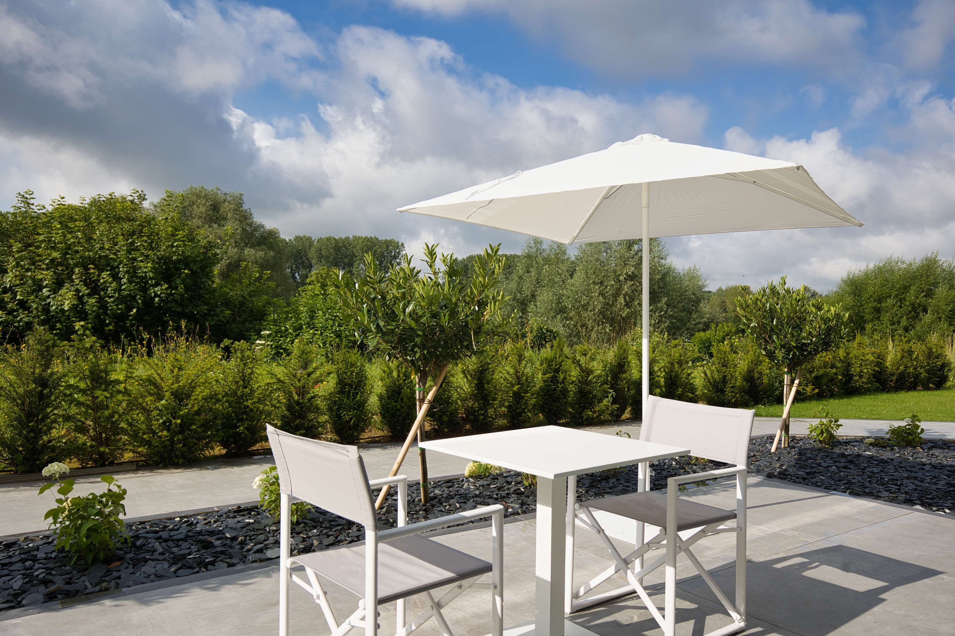 An outdoor patio with a white table and chairs, a white umbrella, and a view of a landscaped garden with trees and shrubs.
