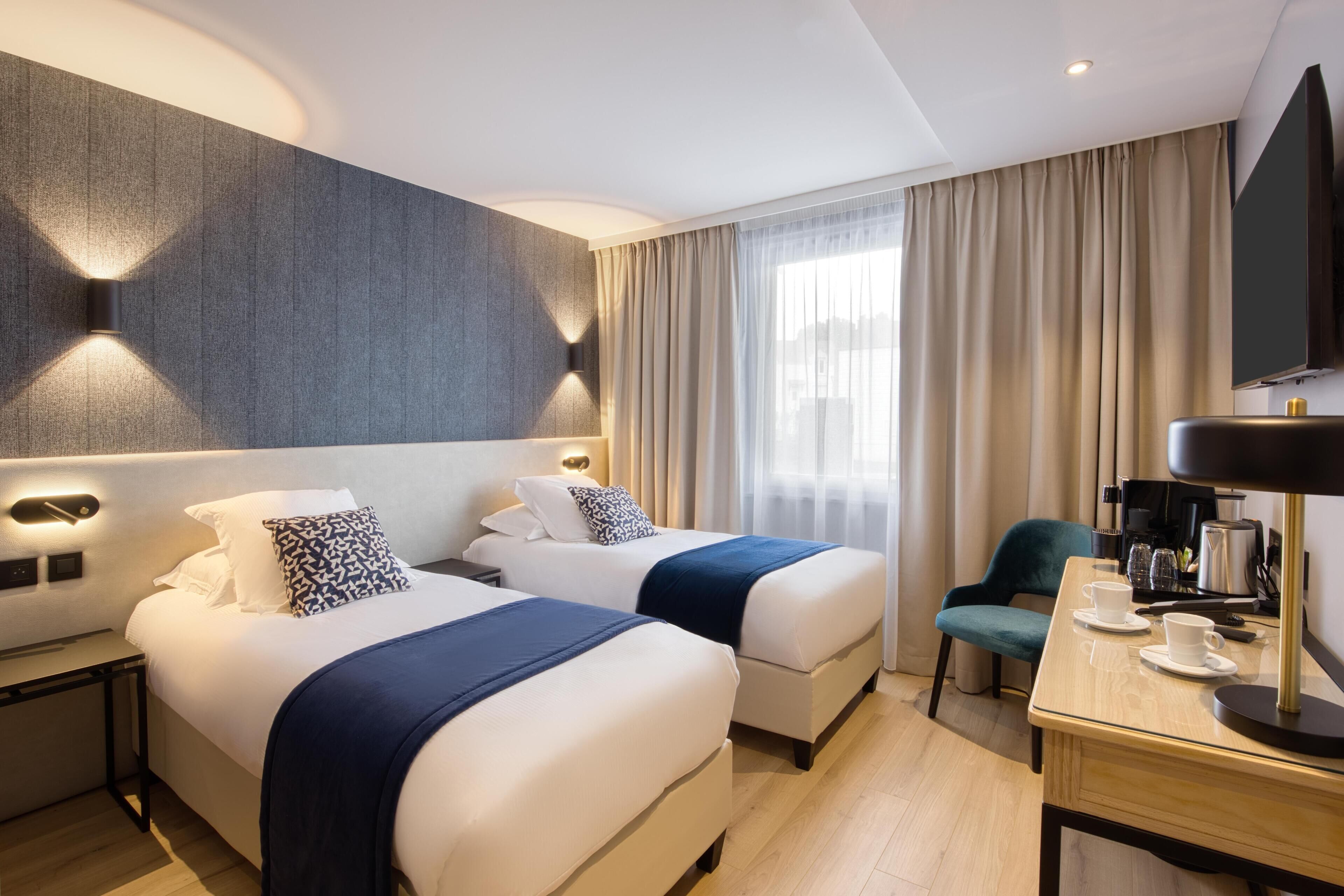 Ein Hotelzimmer mit zwei Betten, einem Schreibtisch mit Kaffeemaschine, einem Fernseher und einem großen Fenster mit Vorhängen.